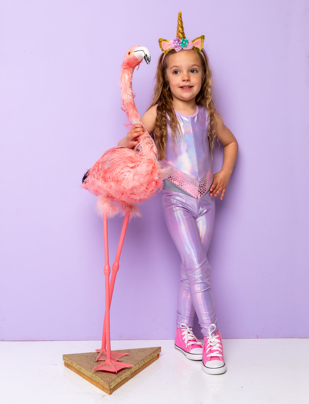 Little girl modelling a lilac holographic onesie or catsuit. She's stood next to a flamingo and has a unicorn horn on her head.