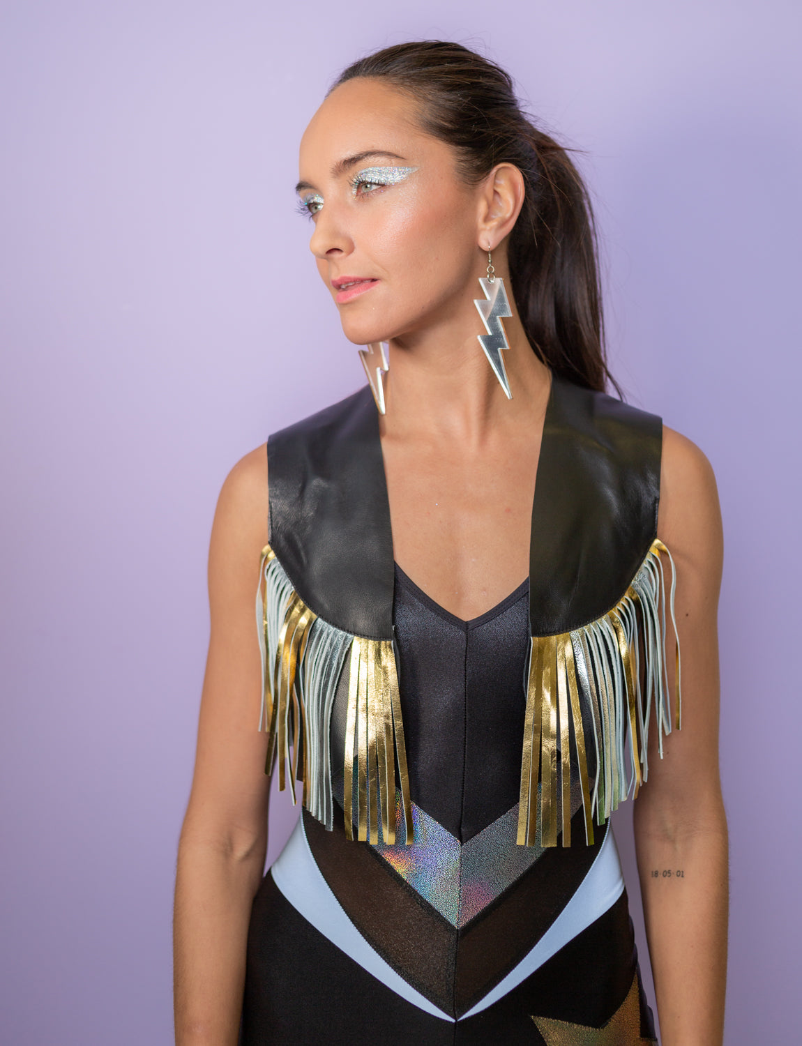 woman wearing a leather waistcoat in black with gold and silver fringing, a black panelled catsuit and perspex lightning bolt earrings