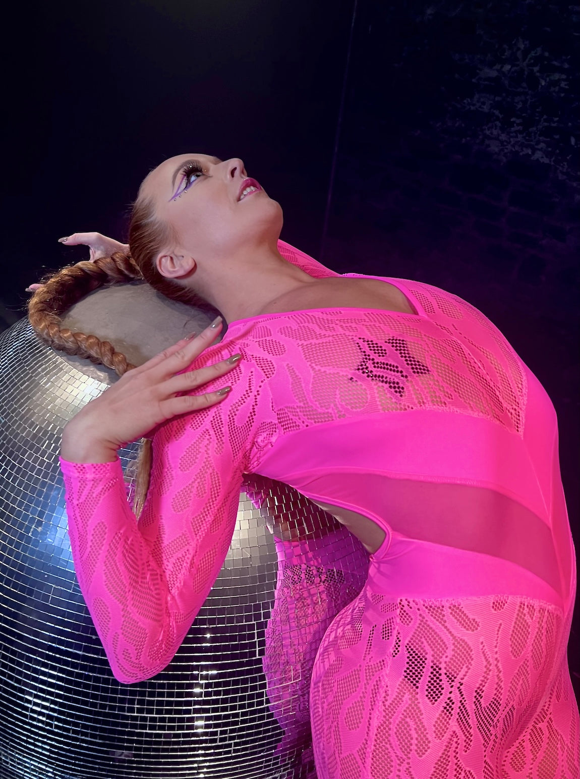 Woman dancer wearing a neon pink mesh flam catsuit leaning back on a giant disco ball.